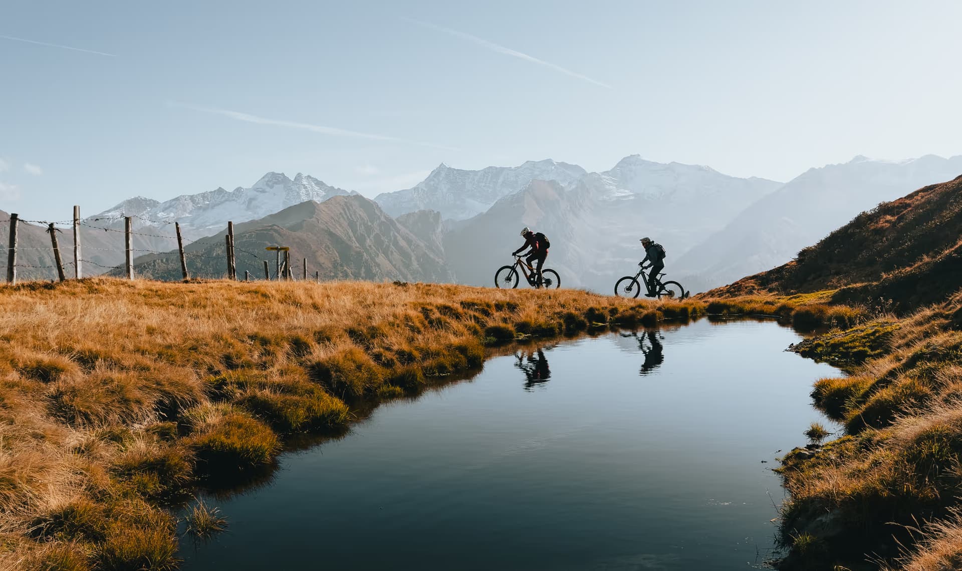 Zwei Mountainbiker auf einem Höhenweg im Herbst mit Bergsee und verschneiten Gipfeln im Wipptal