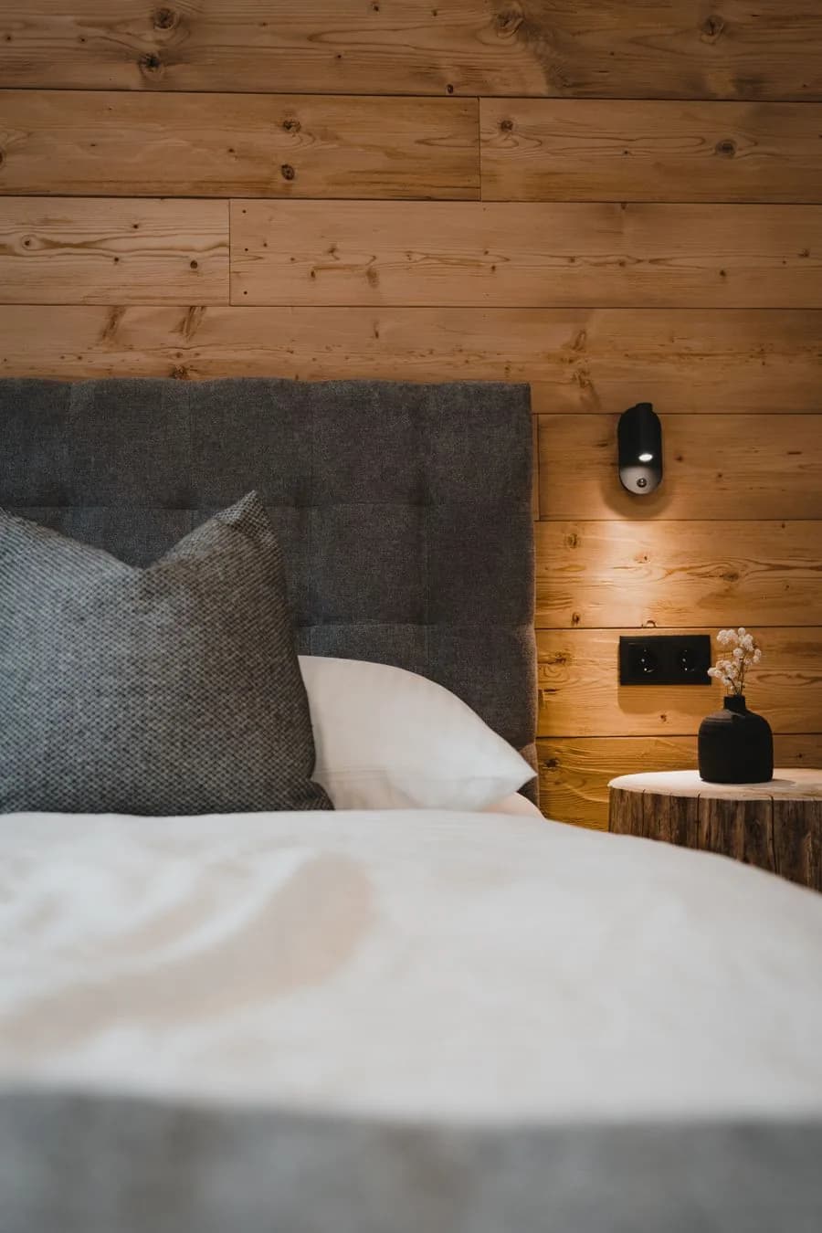 Auszeit apartment Stoacherhof – bedside detail: upholstered headboard, reading lamp on the wooden wall and tree-trunk table with dried flower | holiday flat Wipptal Tyrol